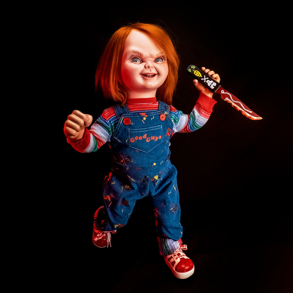 Ultimate Chucky doll.  Ultimate Chucky Doll front view. Red hair, blue eyes, freckles, cleft chin. Face sneering. Wearing red, white, green and blue striped shirt under blue overalls, red buttons, red printed text reads Good Guys on front pocket, red and white sneakers. Action shot, Left hand raised holding knife, right hand raised, clenched in fist.
