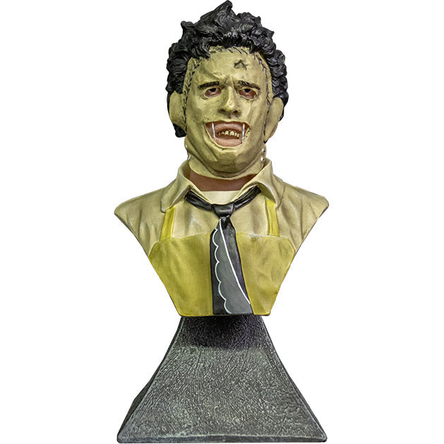 Mini Bust. Front view. Leatherface. Head, shoulders and upper chest of a man wearing a stitched together human face mask, black hair. Wearing a tan shirt with a black and white necktie, yellow apron.  Set on gray stone textured base.