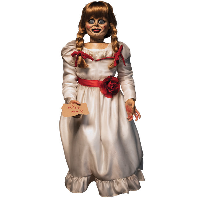 Doll, front view. Large blue eyes, distressed finish on face. Blond hair with bangs, two braids tied with red ribbon. White, floor-length dress with red trim at chest and red belt with rose at waist. Holding card in right hand, red handwritten text reads Miss me? Left hand has blood on palm and fingers.