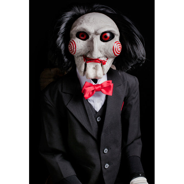 Close up of Saw Billy Puppet prop, above waist. balding with black hair, white face, black-rimmed red eyes, red spirals on cheeks, red lips on hinged ventriloquist dummy mouth. Wearing red bowtie, white collared shirt, black vest and suit coat.