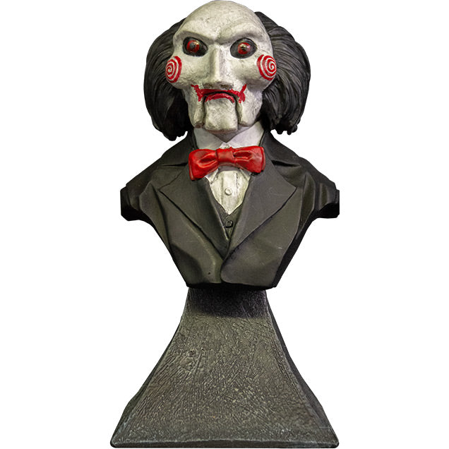  Mini bust, front view. Saw Billy Puppet. Head shoulders and upper chest. Balding with black hair, white face, black-rimmed red eyes, red spirals on cheeks, red lips on hinged ventriloquist dummy mouth. Wearing red bowtie, white collared shirt, black suit coat. Set on gray stone textured base.