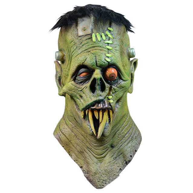 Mask, head, neck and upper chest.  Front view.  Frankenstein-like monster face. Black hair, elongated forehead, flat head, bracket attached on right side, 2 silver posts on either side of head. Green skin, Large wound with light green bandages, on left side of forehead, runs down through left eye and upper lip. Large pointed ears.  Black-rimmed orange eyes, left eye bulging.  No nose, down-turned mouth, long sharp yellowed teeth. 