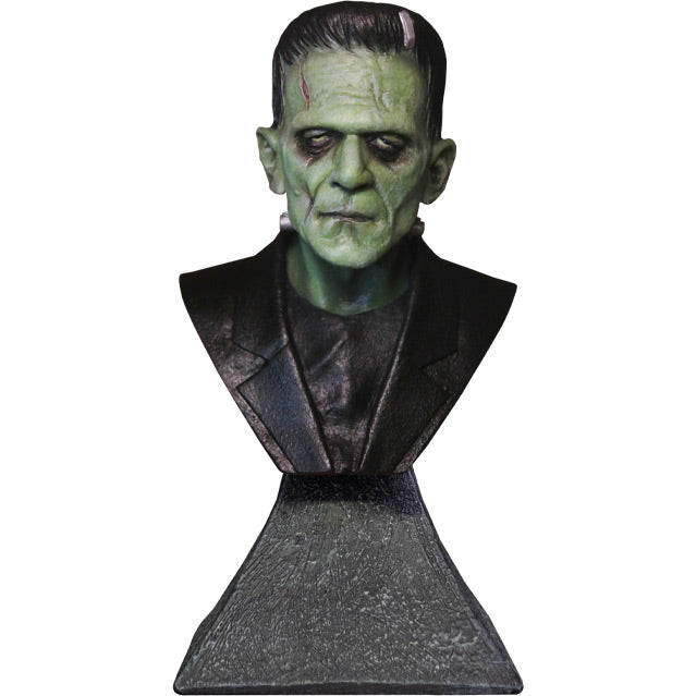 Mini bust, head, neck and chest, front view. Pale greenish skin short black hair, metal bracket on top left forehead, wound on right side of forehead, heavy brow, dark circles around eyes, dark lips, metal bolts on sides of neck.  Black shirt and jacket.