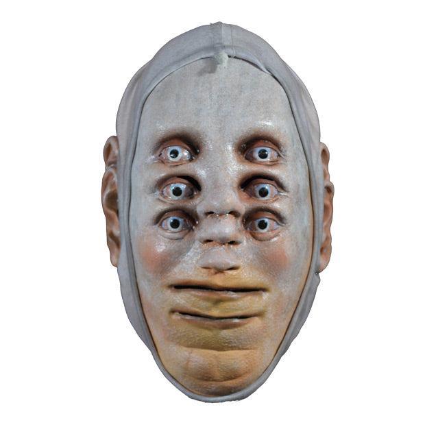 Mask. Front view. Pale white skin. 3 sets of pale blue eyes one above the other, 3 noses one above the other in center of face, two mouths, one above the other at bottom of face, 3 ears on each side of head. White cloth wrap around head to edges of face.