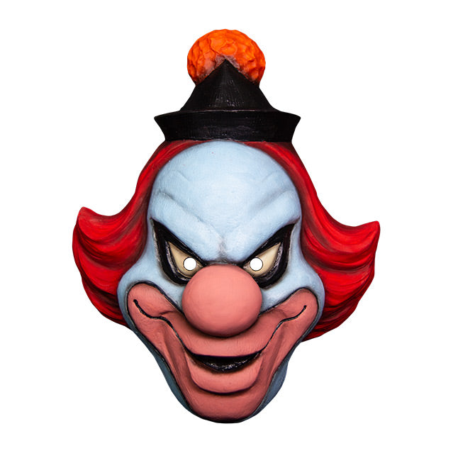 Mask, front view. White clown face, Black-rimmed upturned white eyes, large pale pink nose and lips, menacing smile. Red hair, black cap with orange pompom at top.