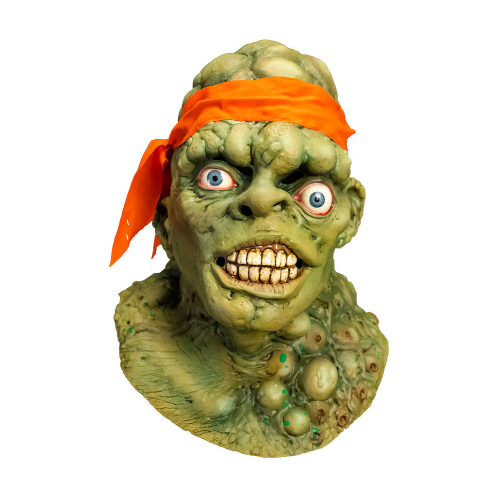 Mask, head, neck and upper chest. Front view. Green lumpy blistered flesh, bald with bright orange headband tied around forehead. Misaligned blue eyes, crooked nose. Lips open showing large dirty teeth. Bumpy neck and chest with sores.