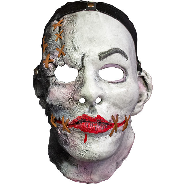Mask, head and neck, front view.  White flesh, black strap across hairline, distressed skin on right side of face, brown stiches on forehead above right eye and on sides of painted on red mouth.  Gray left eyebrow.