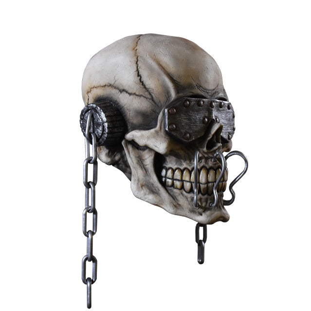 Mask, right side view. Skull wearing metal eye covering with rivets, ear covers with chains hanging off of them, four silver metal hooks attached above to below teeth.