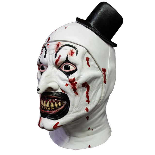 Mask, head and neck, left side view. Evil grinning, black and white clown face with blood spatter, high black painted eyebrows, black around eyes and mouth, black dot on tip of nose, pink gums and yellow teeth. wearing tiny black top hat.