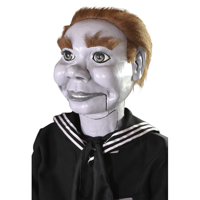 Prop. Close up of face. Ventriloquist dummy, white face and hands, light brown hair and bushy eyebrows, gray eyes, wearing black coveralls, white stripes at neck.