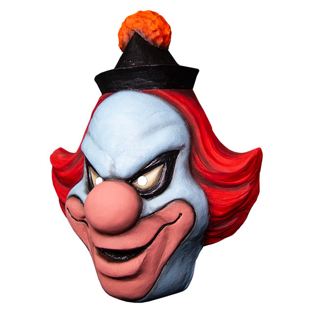 Mask, left side view. White clown face, Black-rimmed upturned white eyes, large pale pink nose and lips, menacing smile. Red hair, black cap with orange pompom at top.