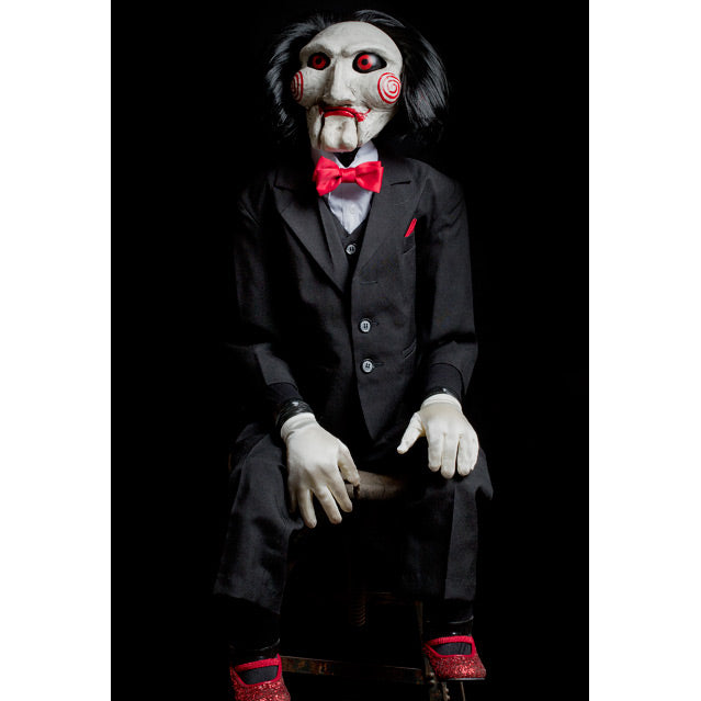  Saw Billy Puppet prop, balding with black hair, white face, black-rimmed red eyes, red spirals on cheeks, red lips on hinged ventriloquist dummy mouth. Wearing red bowtie, white collared shirt, black vest and suit coat, white gloves, black pants and red shoes. Sitting with hands on lap.