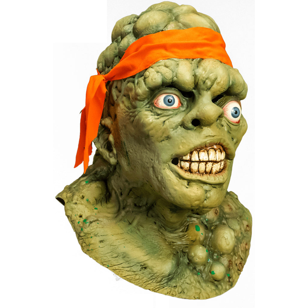 Mask, head, neck and upper chest. right view. Green lumpy blistered flesh, bald with bright orange headband tied around forehead. Misaligned blue eyes, crooked nose. Lips open showing large dirty teeth. Bumpy neck and chest with sores.