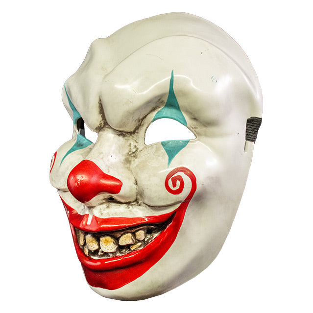 Plastic face mask, left side view. Dirty white clown face, menacing grin, blue painted triangles above and below eyes, red on nose, large red painted smile with spirals on cheeks, dirty yellow teeth.