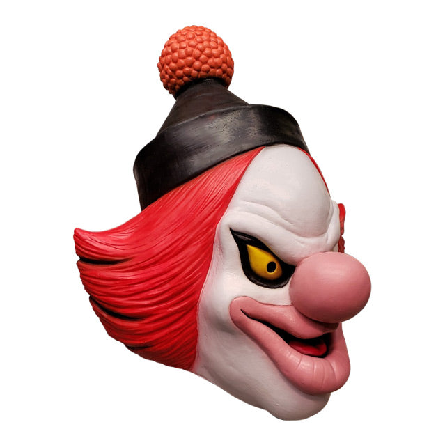 Mask, right side view. White clown face, Black-rimmed upturned yellow eyes, large pale pink nose and lips, menacing smile with mouth slightly open, showing red tongue. Red hair, black cap with orange pompom at top.