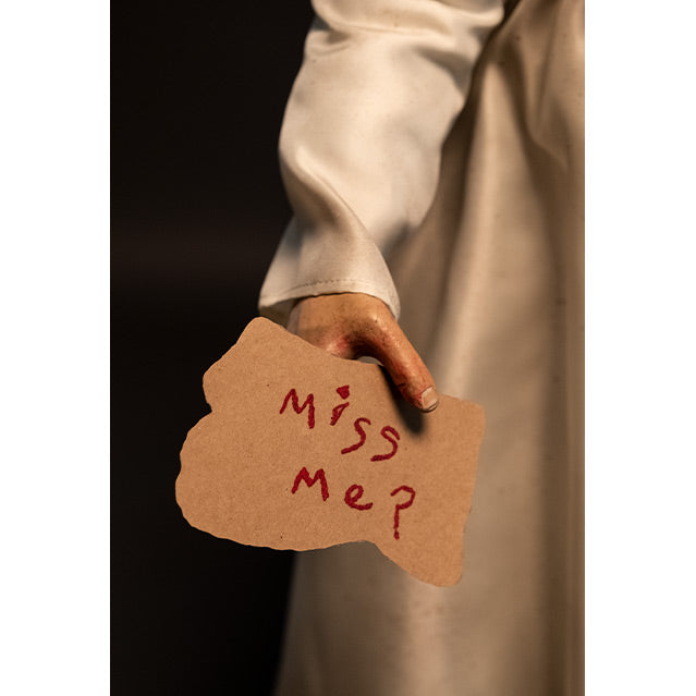 Close up of doll right hand, black background.  White dress, holding card, red handwritten text reads Miss me?