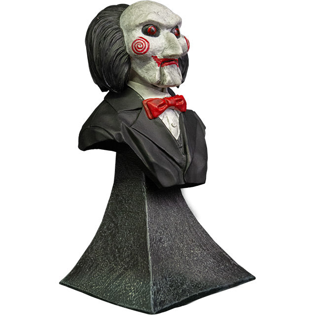 Mini bust, right view. Saw Billy Puppet. Head shoulders and upper chest. Balding with black hair, white face, black-rimmed red eyes, red spirals on cheeks, red lips on hinged ventriloquist dummy mouth. Wearing red bowtie, white collared shirt, black suit coat. Set on gray stone textured base.