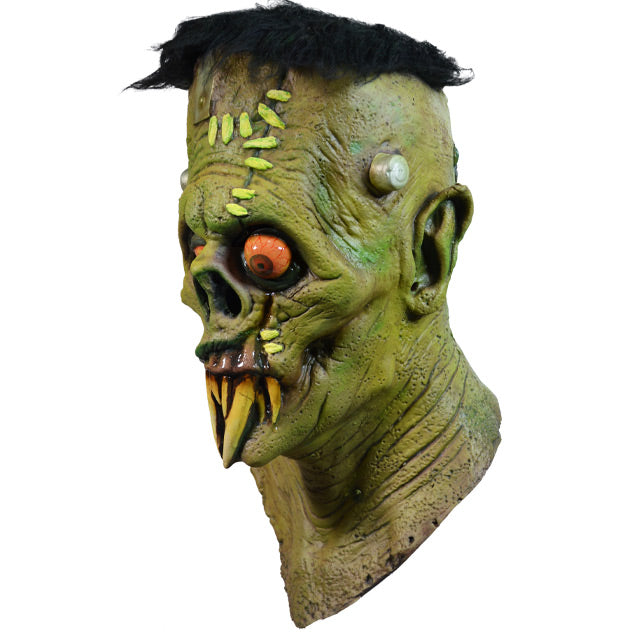 Mask, head, neck and upper chest.  Left view.  Frankenstein-like monster face. Black hair, elongated forehead, flat head, bracket attached on right side, 2 silver posts on either side of head. Green skin, Large wound with light green bandages, on left side of forehead, runs down through left eye and upper lip. Large pointed ears. Black-rimmed orange eyes, left eye bulging. No nose, down-turned mouth, long sharp yellowed teeth.