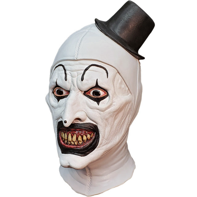 Mask, head and neck, left side view. Evil grinning, black and white clown face, high black painted eyebrows, black around eyes and mouth, black dot on tip of nose, pink gums and yellow teeth. wearing tiny black top hat.