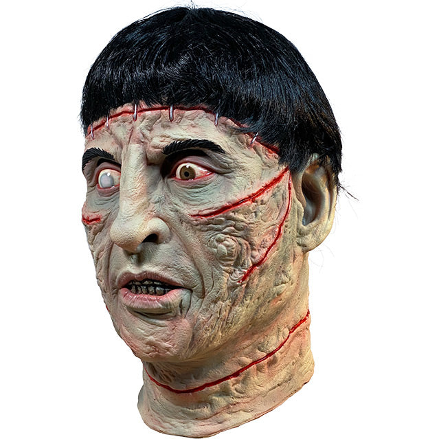 Mask, left side  view, head and neck. Short black hair, staples in forehead, red wounds below eyes, on cheeks and on neck, red-rimmed eyes, right eye iris is white, left eye iris is brown. Mouth slightly open, showing teeth.