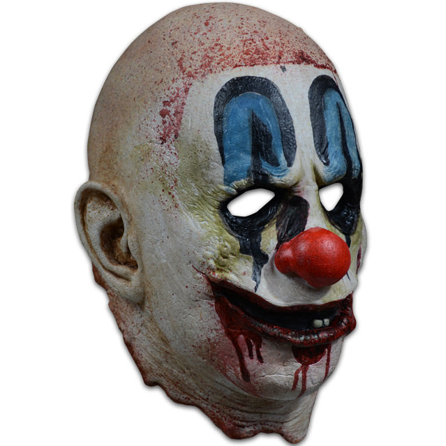 Mask, right side view. Bald head, blood spattered, white, dirty clown makeup. Blue painted above black-rimmed eyes, large red clown nose, red clown mouth open with 2 teeth showing, blood dripping from mouth.