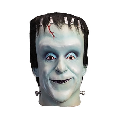 Mask, head and neck, front view. Herman Munster, white-blue skin, tall forehead. Black hair on head, silver brackets across top of forehead, small red cut on right side. No eyebrows, blue eyelids, brown eyes. Mouth closed, smiling, black lips. Metal posts on side of neck.
