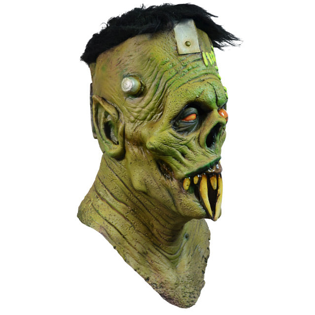Mask, head, neck and upper chest. Right view.  Frankenstein-like monster face. Black hair, elongated forehead, flat head, bracket attached on right side, 2 silver posts on either side of head. Green skin, Large wound with light green bandages, on left side of forehead, runs down through left eye and upper lip. Large pointed ears. Black-rimmed orange eyes, left eye bulging. No nose, down-turned mouth, long sharp yellowed teeth.