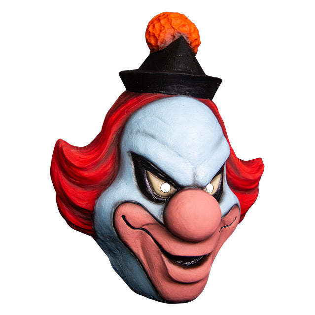 Mask, right side view. White clown face, Black-rimmed upturned white eyes, large pale pink nose and lips, menacing smile. Red hair, black cap with orange pompom at top.