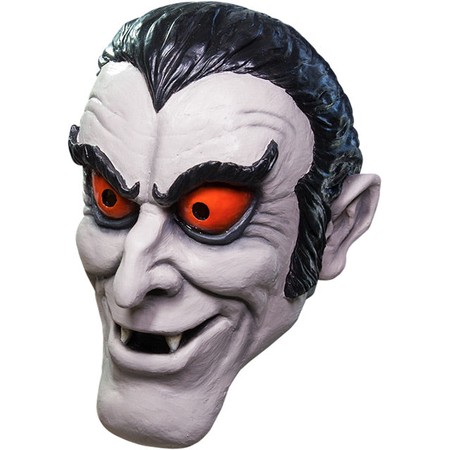 Mask, left side view. Pale gray cartoon vampire face. black hair with widow's peak, upturned black eyebrows, red-orange eyes, pointed ears, mouth open showing fangs.