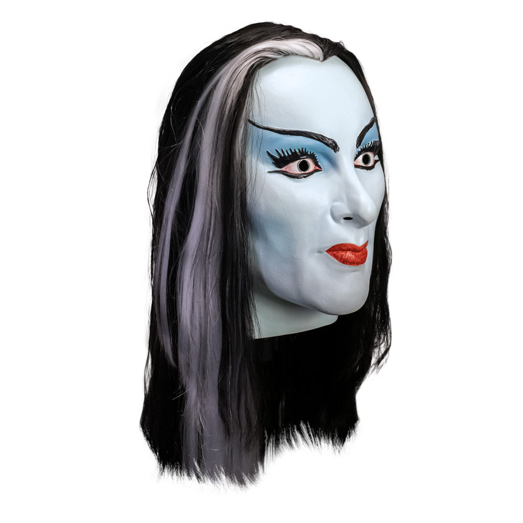 Mask, head and neck, right side view. Lily Munster, white-blue skin. Black hair with white streak in middle of forehead. Thin black eyebrows, blue eyelids, big eyelashes, black winged eyeliner. Mouth closed, smiling, bright red lips.
