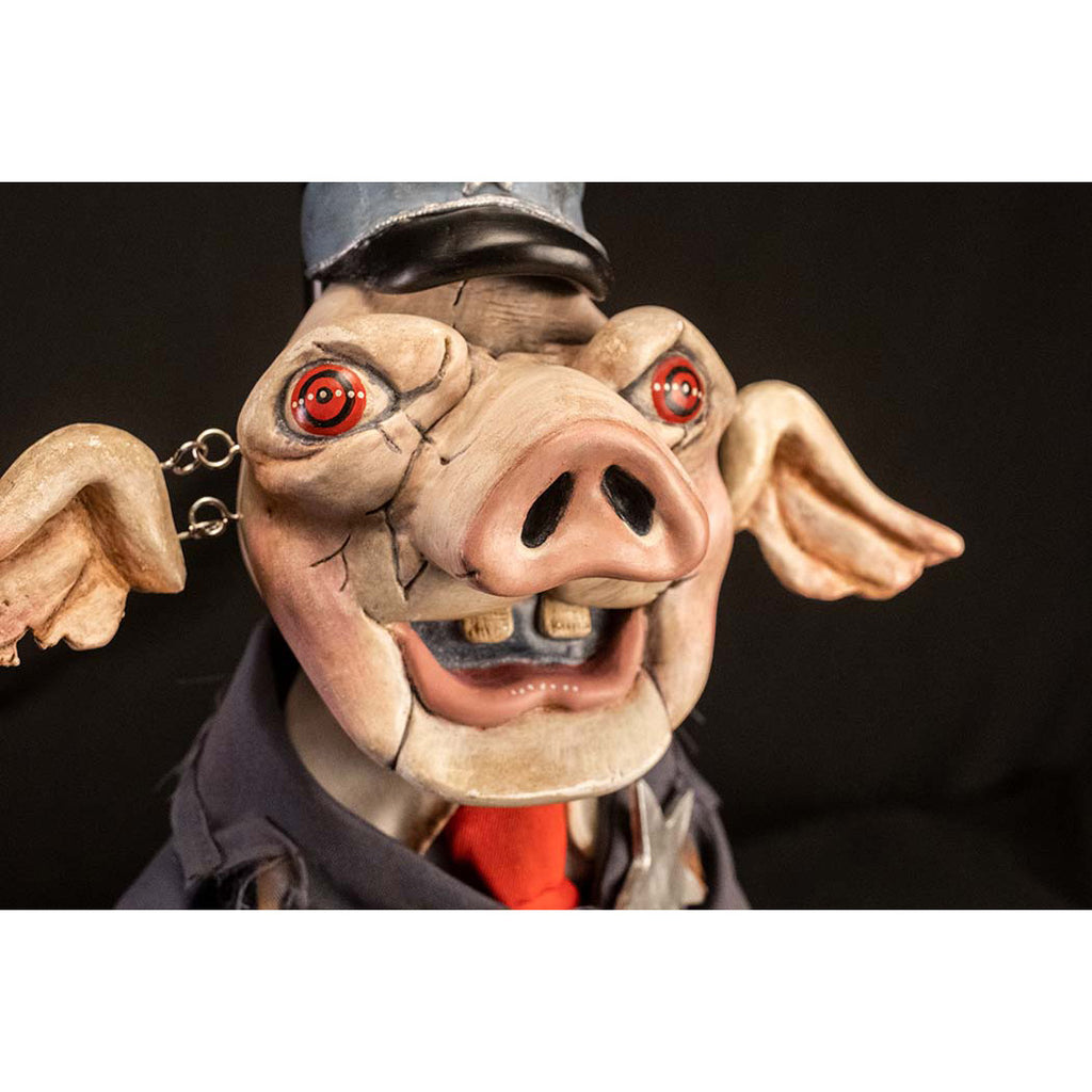 Mr. Snuggles puppet prop. Black background. Close up of pink pig face, red eyes, open mouth with two buck teeth, ears attached with metal links, blue hat, black brim. Wearing distressed blue suit, with silver star, dirty white shirt, red necktie.