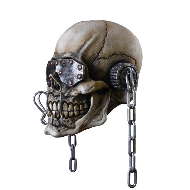 Mask, left side view. Skull wearing metal eye covering with rivets, ear covers with chains hanging off of them, four silver metal hooks attached above to below teeth.