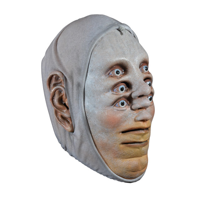 Mask. Right side view. Pale white skin. 3 sets of pale blue eyes one above the other, 3 noses one above the other in center of face, two mouths, one above the other at bottom of face, 3 ears on each side of head. White cloth wrap around head to edges of face.