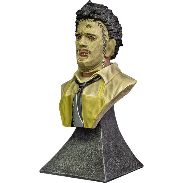 Mini Bust. left view. Leatherface. Head, shoulders and upper chest of a man wearing a stitched together human face mask, black hair. Wearing a tan shirt with a black and white necktie, yellow apron. Set on gray stone textured base.