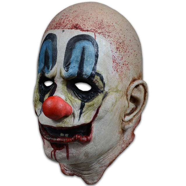 Mask, left side view. Bald head, blood spattered, white, dirty clown makeup. Blue painted above black-rimmed eyes, large red clown nose, red clown mouth open with 2 teeth showing, blood dripping from mouth.