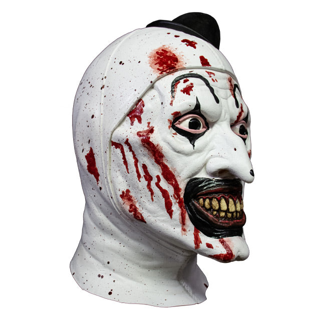 Mask, head and neck, right side view. Evil grinning, black and white clown face with blood spatter, high black painted eyebrows, black around eyes and mouth, black dot on tip of nose, pink gums and yellow teeth. wearing tiny black top hat.