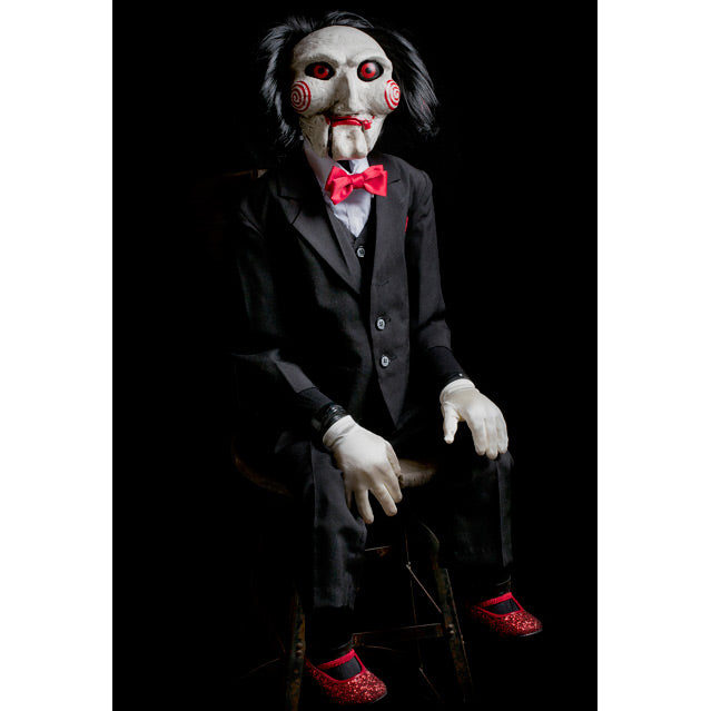 Saw Billy Puppet prop, balding with black hair, white face, black-rimmed red eyes, red spirals on cheeks, red lips on hinged ventriloquist dummy mouth. Wearing red bowtie, white collared shirt, black vest and suit coat, white gloves, black pants and red shoes. Sitting with hands on lap.