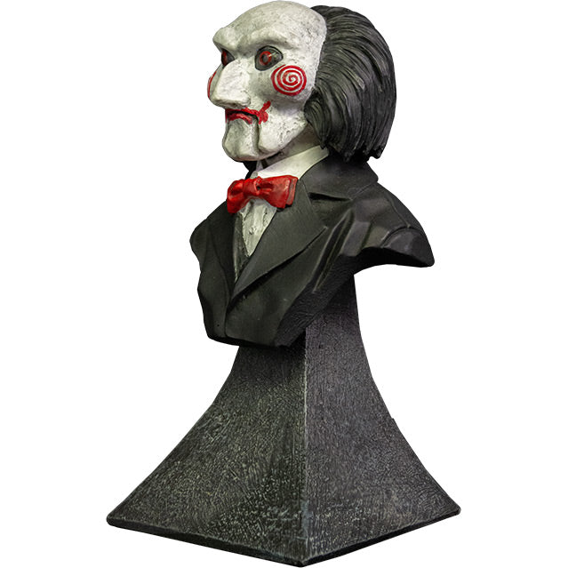 Mini bust, left view. Saw Billy Puppet. Head shoulders and upper chest. Balding with black hair, white face, black-rimmed red eyes, red spirals on cheeks, red lips on hinged ventriloquist dummy mouth. Wearing red bowtie, white collared shirt, black suit coat. Set on gray stone textured base.