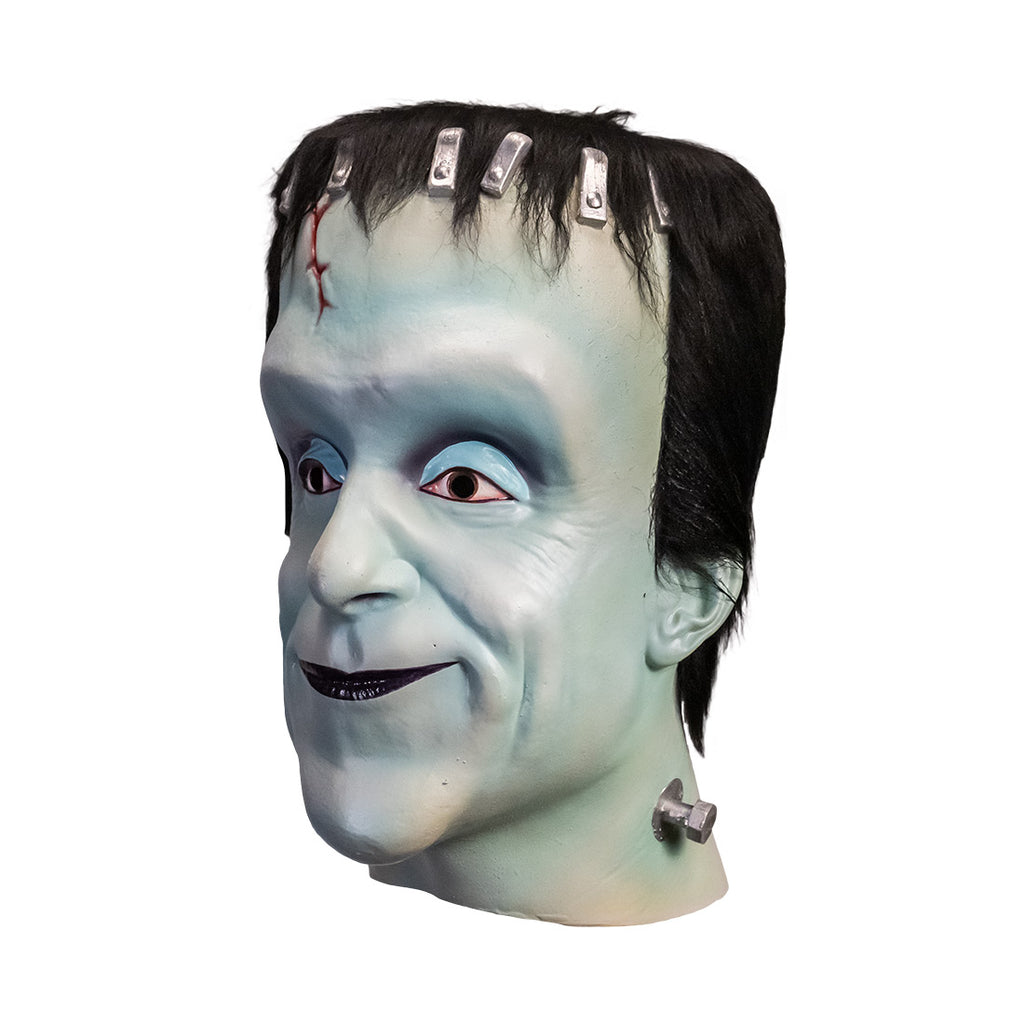 Mask, head and neck, left side view. Herman Munster, white-blue skin, tall forehead. Black hair on head, silver brackets across top of forehead, small red cut on right side. No eyebrows, blue eyelids, brown eyes. Mouth closed, smiling, black lips. Metal posts on side of neck.