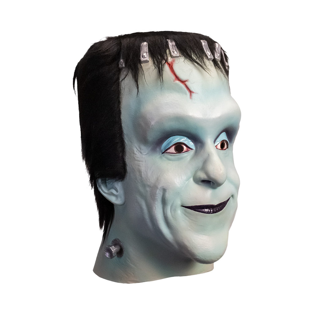 Mask, head and neck, right side view. Herman Munster, white-blue skin, tall forehead. Black hair on head, silver brackets across top of forehead, small red cut on right side. No eyebrows, blue eyelids, brown eyes. Mouth closed, smiling, black lips. Metal posts on side of neck.
