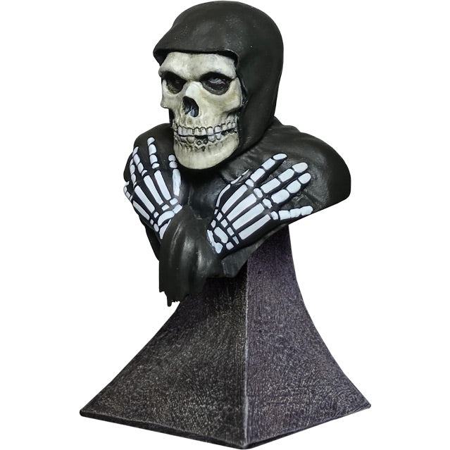 Mini bust, left side view. Head, shoulders and upper chest. Misfits Fiend, skeleton face and hands, crossed, wearing black hooded cloak. Set on gray stone textured base.