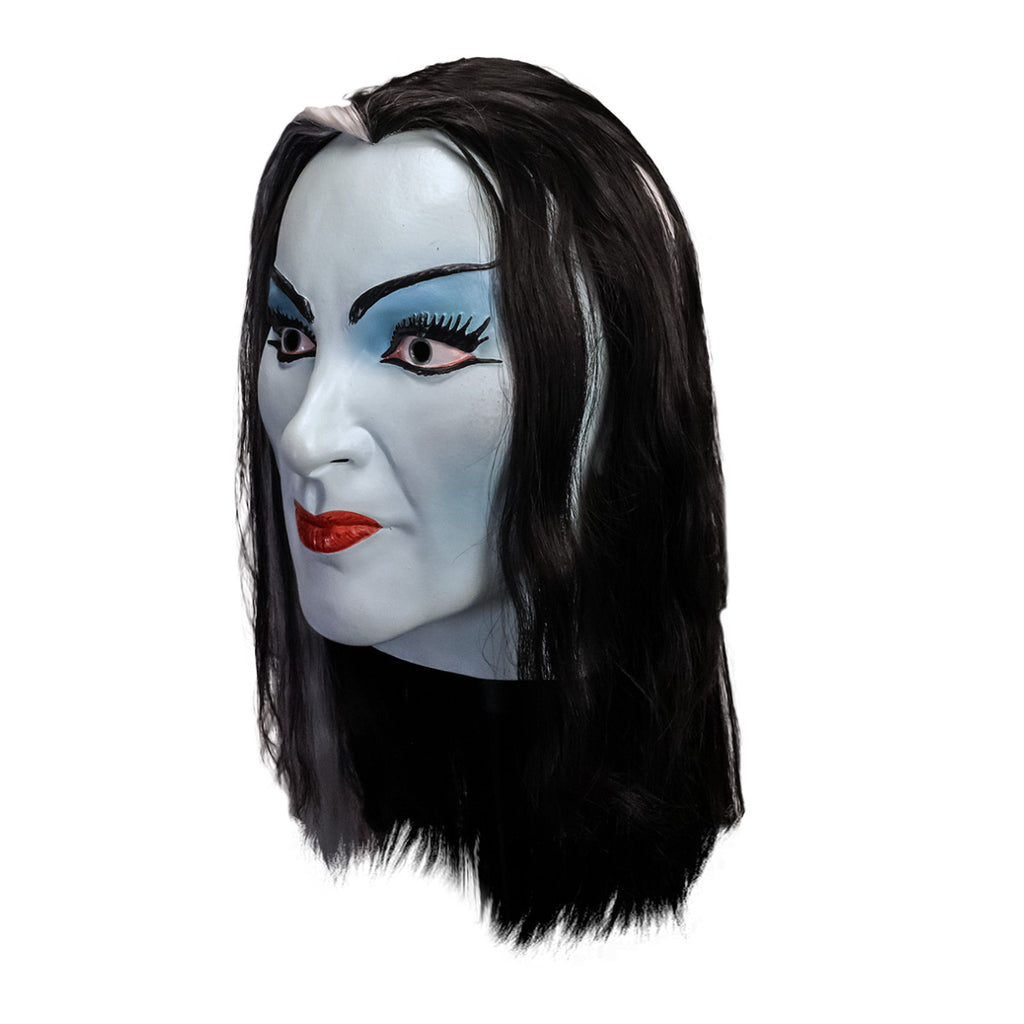 Mask, head and neck, left side view. Lily Munster, white-blue skin. Black hair with white streak in middle of forehead. Thin black eyebrows, blue eyelids, big eyelashes, black winged eyeliner. Mouth closed, smiling, bright red lips.