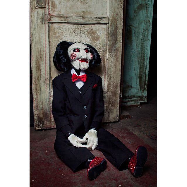 Saw Billy Puppet prop, balding with black hair, white face, black-rimmed red eyes, red spirals on cheeks, red lips on hinged ventriloquist dummy mouth. Wearing red bowtie, white collared shirt, black vest and suit coat, white gloves, black pants and red shoes. Sitting on the floor with hands on lap.