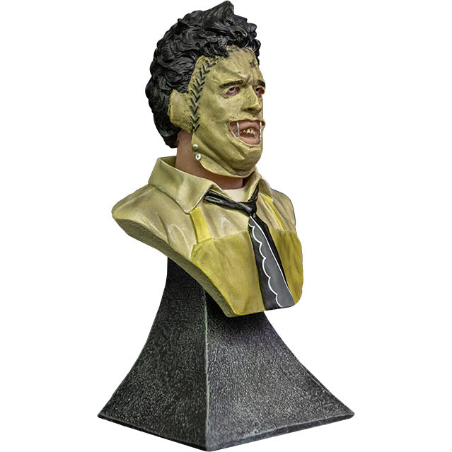 Mini Bust. Right view. Leatherface. Head, shoulders and upper chest of a man wearing a stitched together human face mask, black hair. Wearing a tan shirt with a black and white necktie, yellow apron. Set on gray stone textured base.