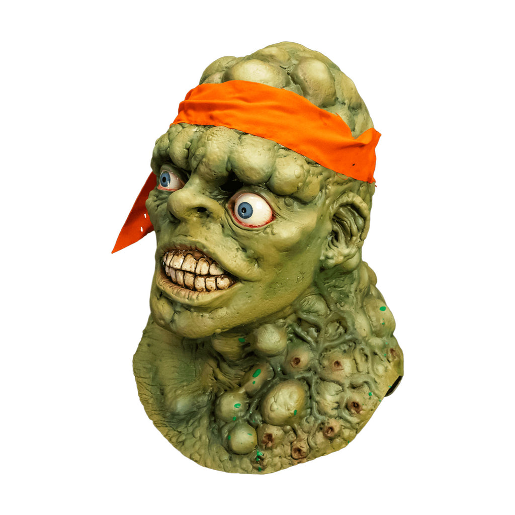 Mask, head, neck and upper chest. Left view. Green lumpy blistered flesh, bald with bright orange headband tied around forehead. Misaligned blue eyes, crooked nose. Lips open showing large dirty teeth. Bumpy neck and chest with sores.