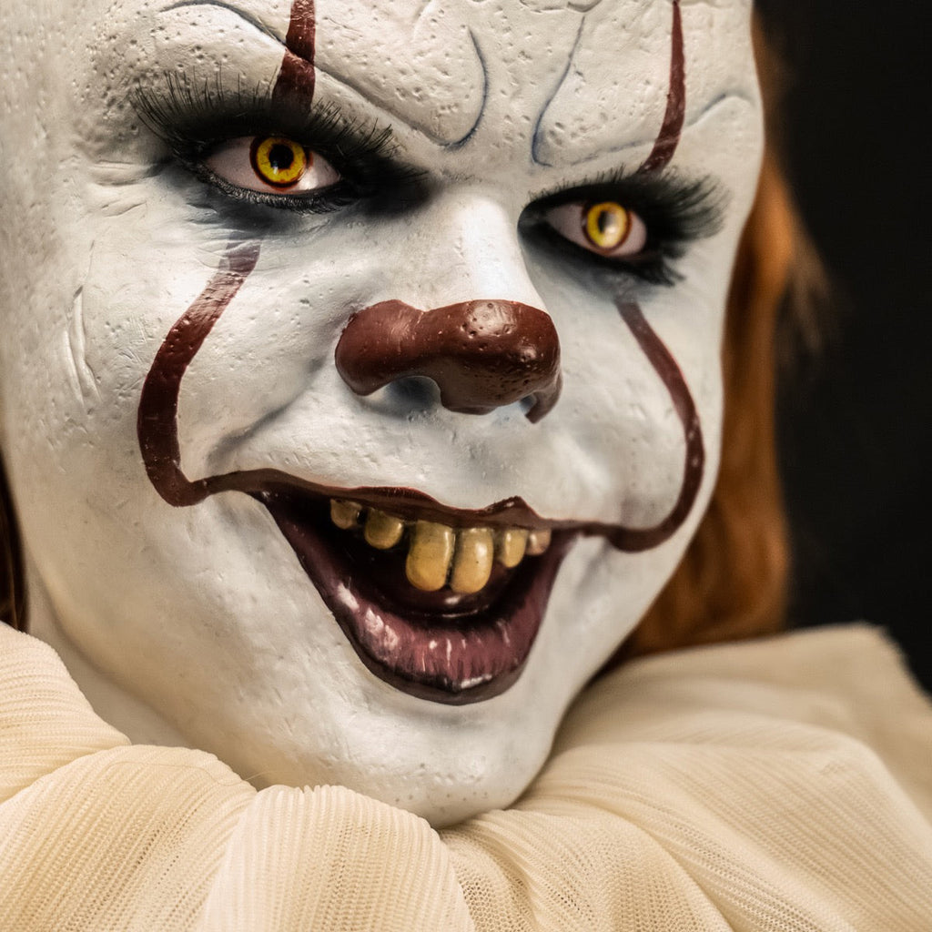 Pennywise doll, close up view. clown face, red hair, white skin, large forehead, yellow eyes and nose, dark lips, creepy smile with crooked yellow buck teeth