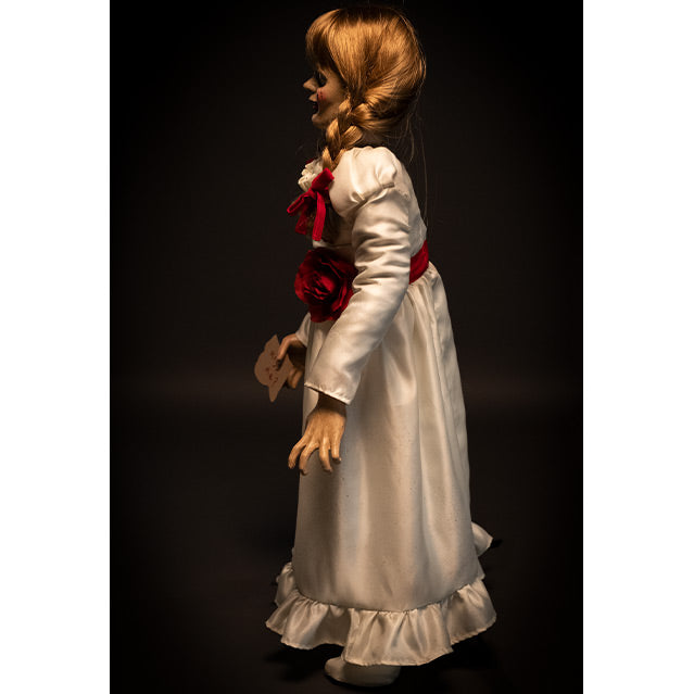 Doll, right side view, black background. Blond hair with bangs, two braids tied with red ribbon. White, floor-length dress with red trim at chest and red belt with rose at waist. Holding card in right hand.