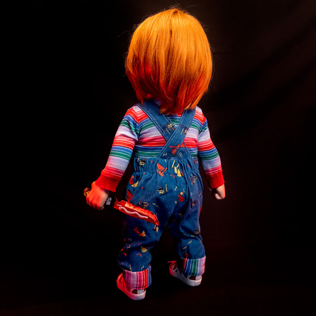 Ultimate Chucky doll, back view.  Red hair, wearing red, white, green and blue striped shirt under blue overalls, Voodoo knife in left hand.