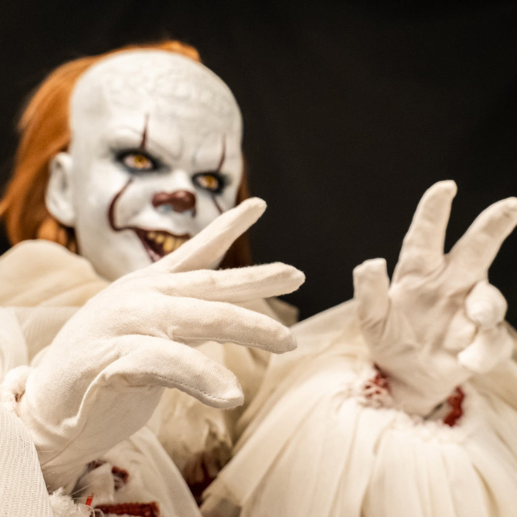 Pennywise doll, close up view of white gloved hands. Background, clown face, red hair, white skin, large forehead, yellow eyes and nose, dark lips, creepy smile with crooked yellow buck teeth
