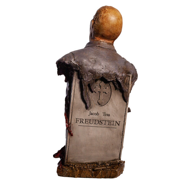 Zombie bust, back view, head, shoulders and chest. Orange-hued shriveled flesh, wearing filthy and gory blue doctor's coat. Gore coming from bottom of torso. Gray tombstone in back, cross in circle at top, text reads Jacob Tess Freudstein, set on wood look base.